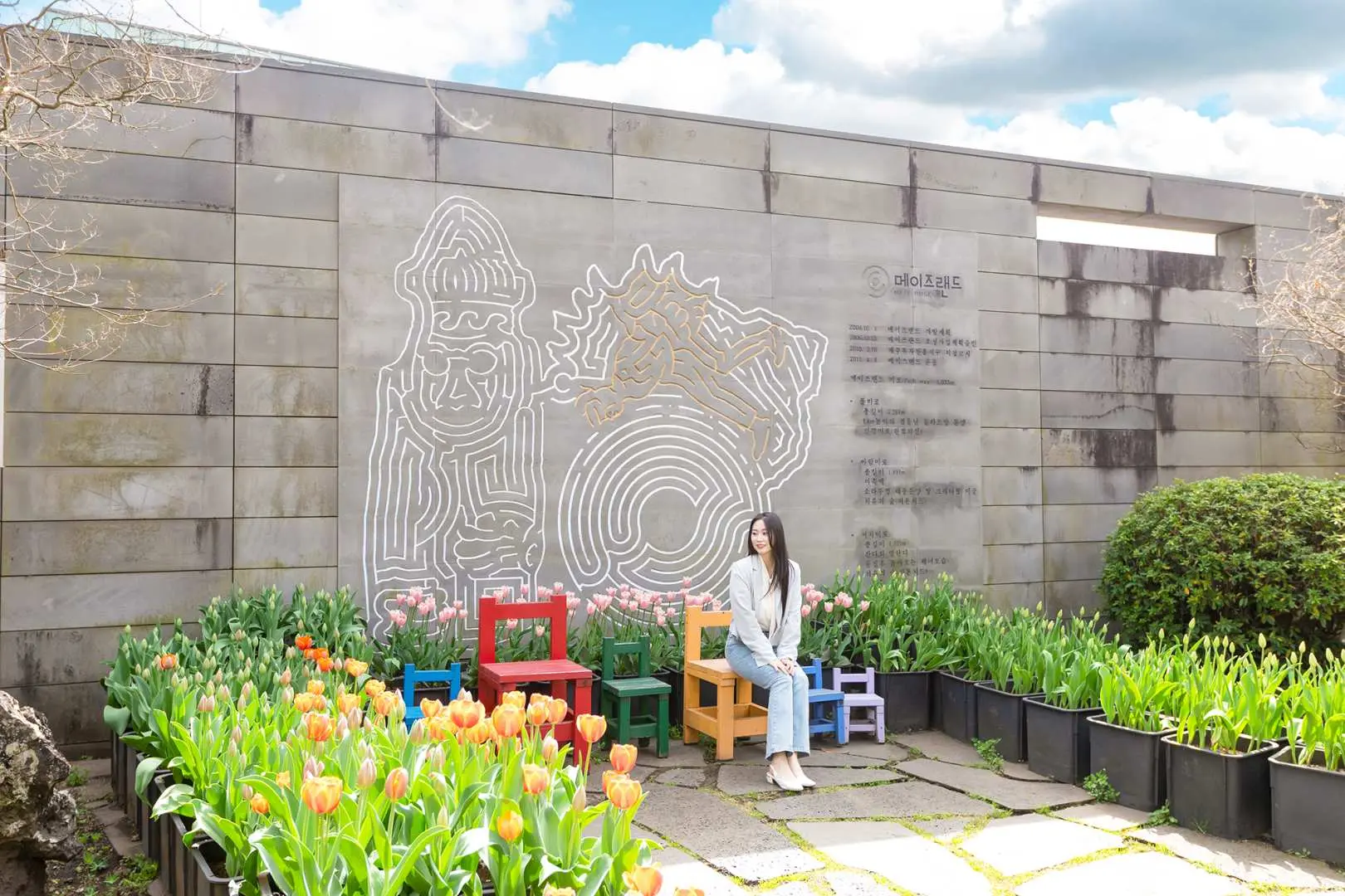 韓國濟州Maze Land迷宮博物館外牆，遊客坐在彩色椅子上，身旁擺滿鬱金香，背景為濟州特色石雕壁畫，充滿藝術氣息。