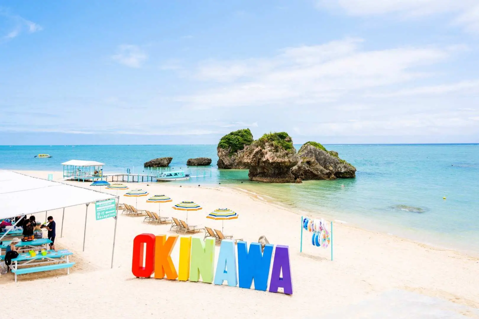 Picturesque Okinawa Shinbara Beach with a vibrant 'OKINAWA' sign on white sands, under a clear blue sky, featuring unique rock formations in the distance.