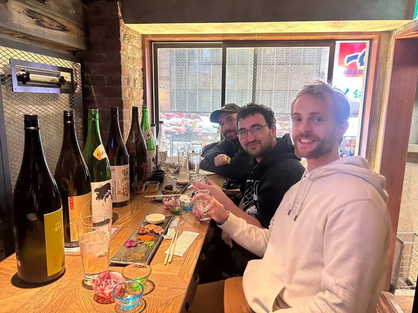 Smiling customers at a bar counter in Shinjuku, Tokyo, enjoying a Japanese sake tasting experience with various bottles and small appetizer dishes visible.