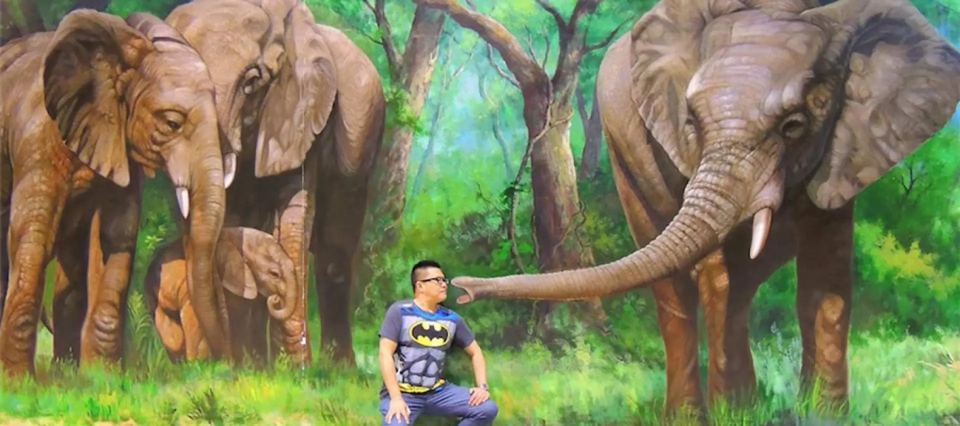 一名男子在芭提雅3D美术馆与逼真的丛林大象3D画作互动，一头大象的长鼻子仿佛正伸向他，营造身临其境的趣味拍照体验。