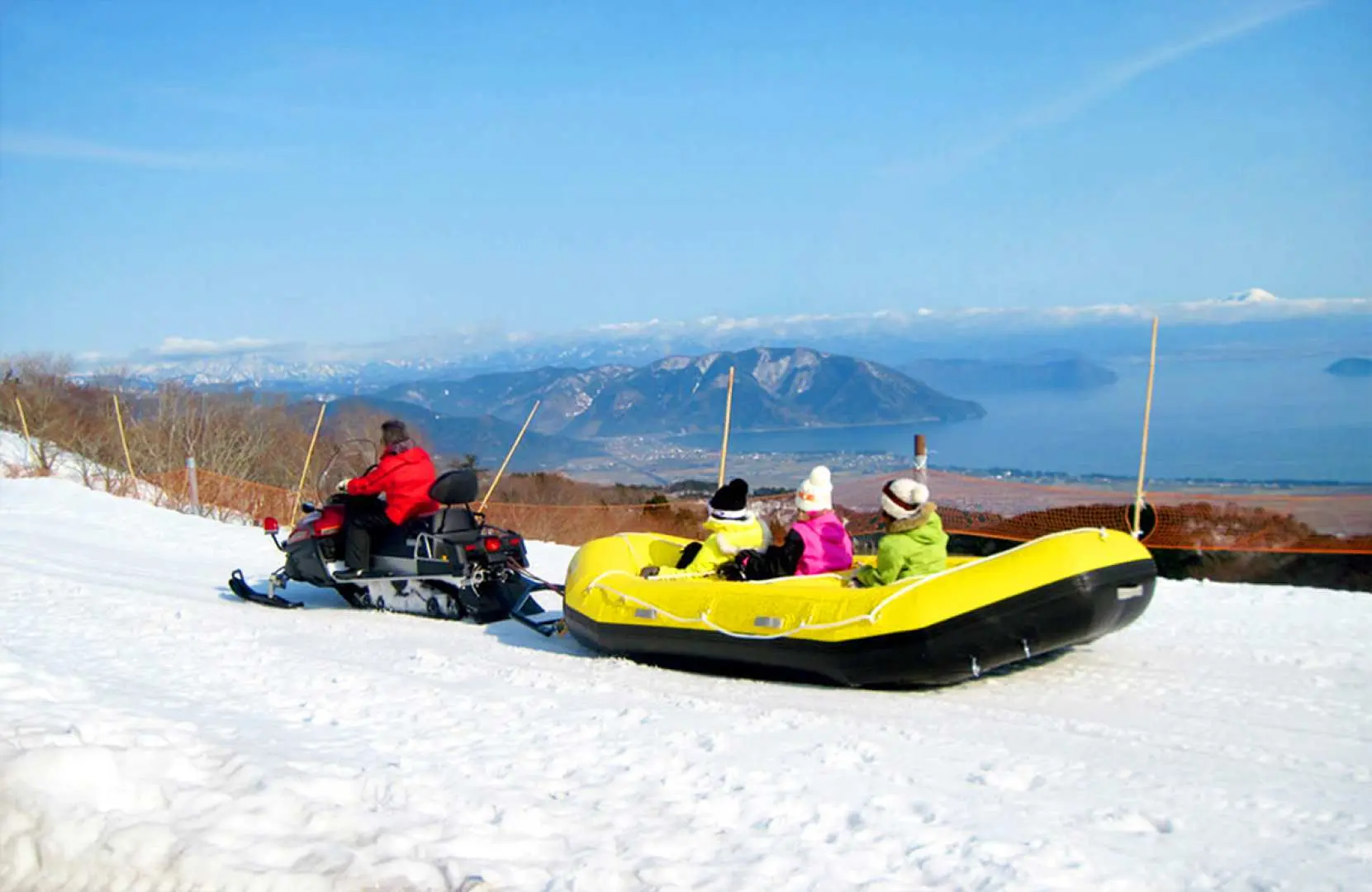 日本の箱館山スキー場でのスリリングなスノーラフティングアクティビティです。スノーモービルが3人の乗客を乗せたインフレータブルラフトを牽引し、雪面を疾走します。背景には琵琶湖と遠くの山々が広がり、スピードと絶景を楽しめます。