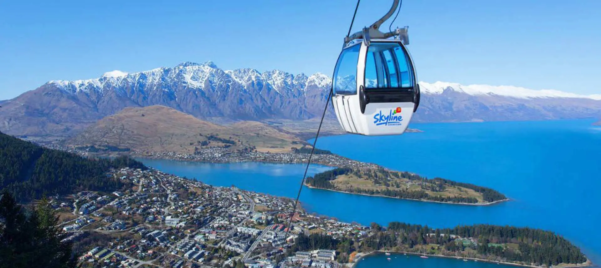 A blue Skyline gondola cabin ascends above Queenstown, offering aerial views of the vibrant town, serene Lake Wakatipu, and surrounding mountains.