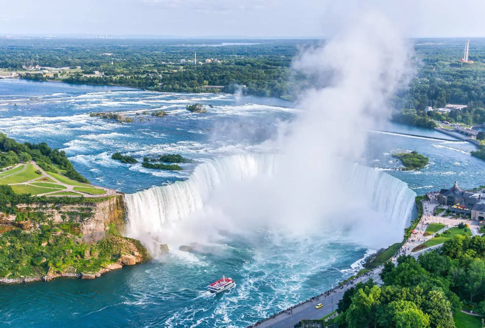 A vast aerial shot of Canada's Niagara Falls showcasing the majestic curve of Horseshoe Falls, American Falls, and Bridal Veil Falls, with a Hornblower Cruise boat navigating the waters.