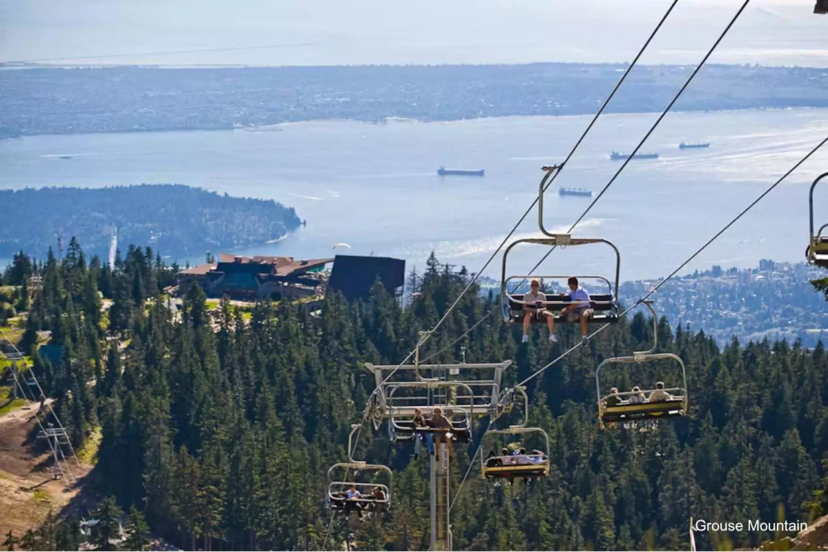 Visitors on a Grouse Mountain chairlift ascend over green treetops, enjoying expansive panoramic views of Vancouver's city, coastline, and the sparkling ocean below.
