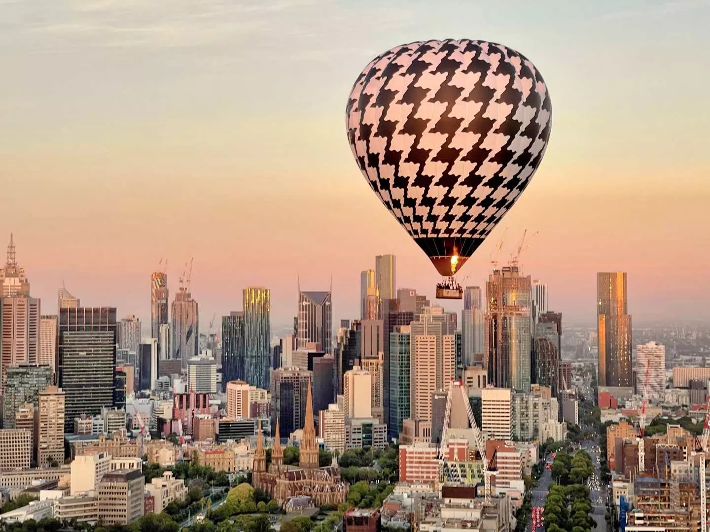 A distinctive black and white patterned hot air balloon soaring majestically above the bustling Melbourne city center and its architectural marvels at dawn.