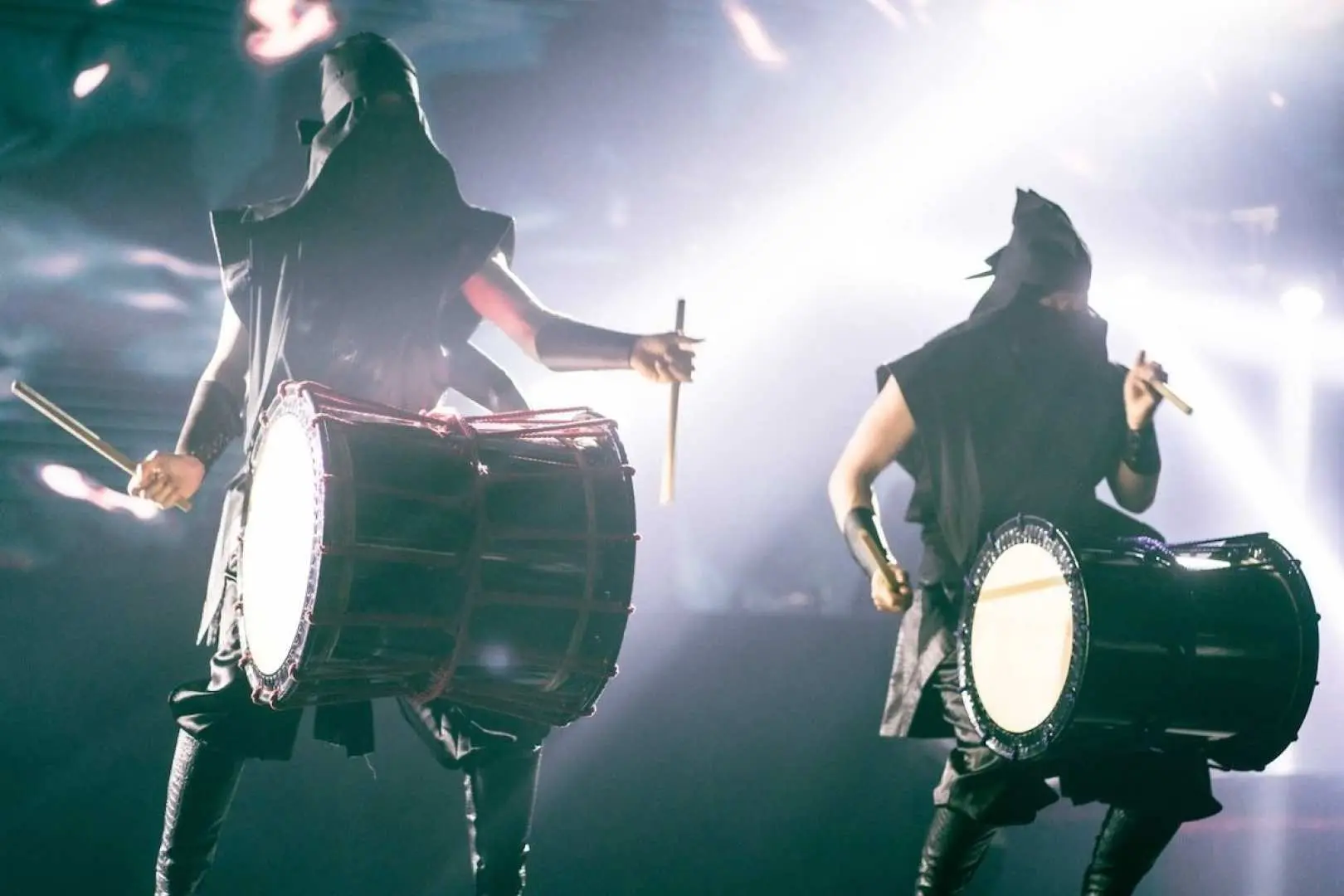 Two powerful performers on stage at the Tokyo Shinjuku Ninja+Kabuki show, seen from behind, intensely playing large traditional Japanese taiko drums under bright lights, creating a dynamic and rhythmic cultural spectacle.