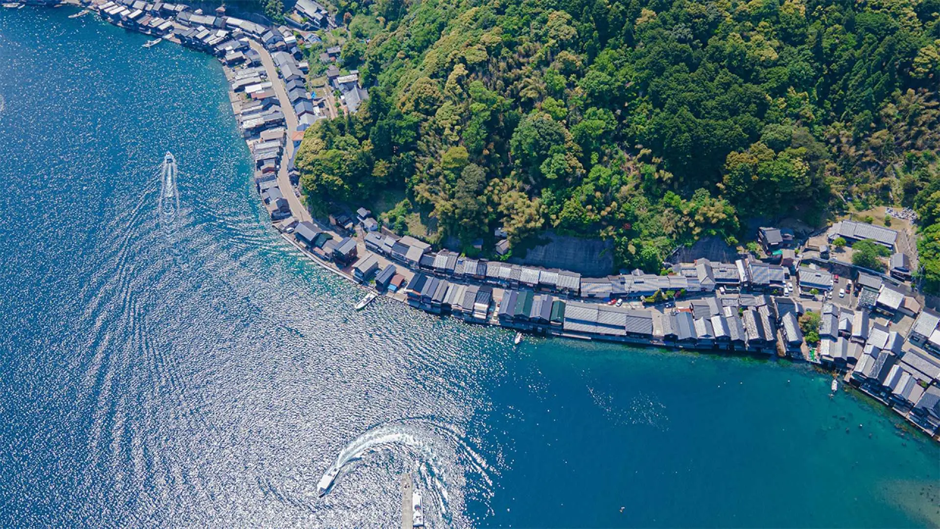 伊根の舟屋群の鳥瞰図。伝統的な木造家屋が曲がりくねった海岸線に沿って並び、緑豊かな山林に囲まれ、紺碧の海を遊覧船が航行する、独特の湾の景観が広がっています。