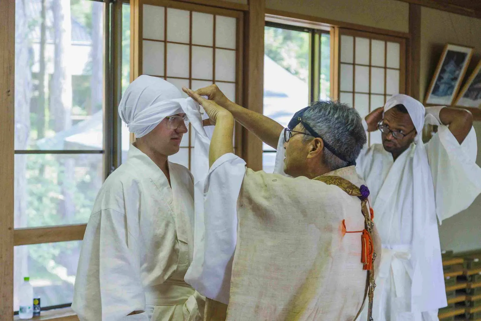 During the Jionji Temple spiritual culture tour in Yamagata, a Yamabushi guide helps a participant don a white head covering, preparing for a Shugendo experience and showcasing traditional rituals.