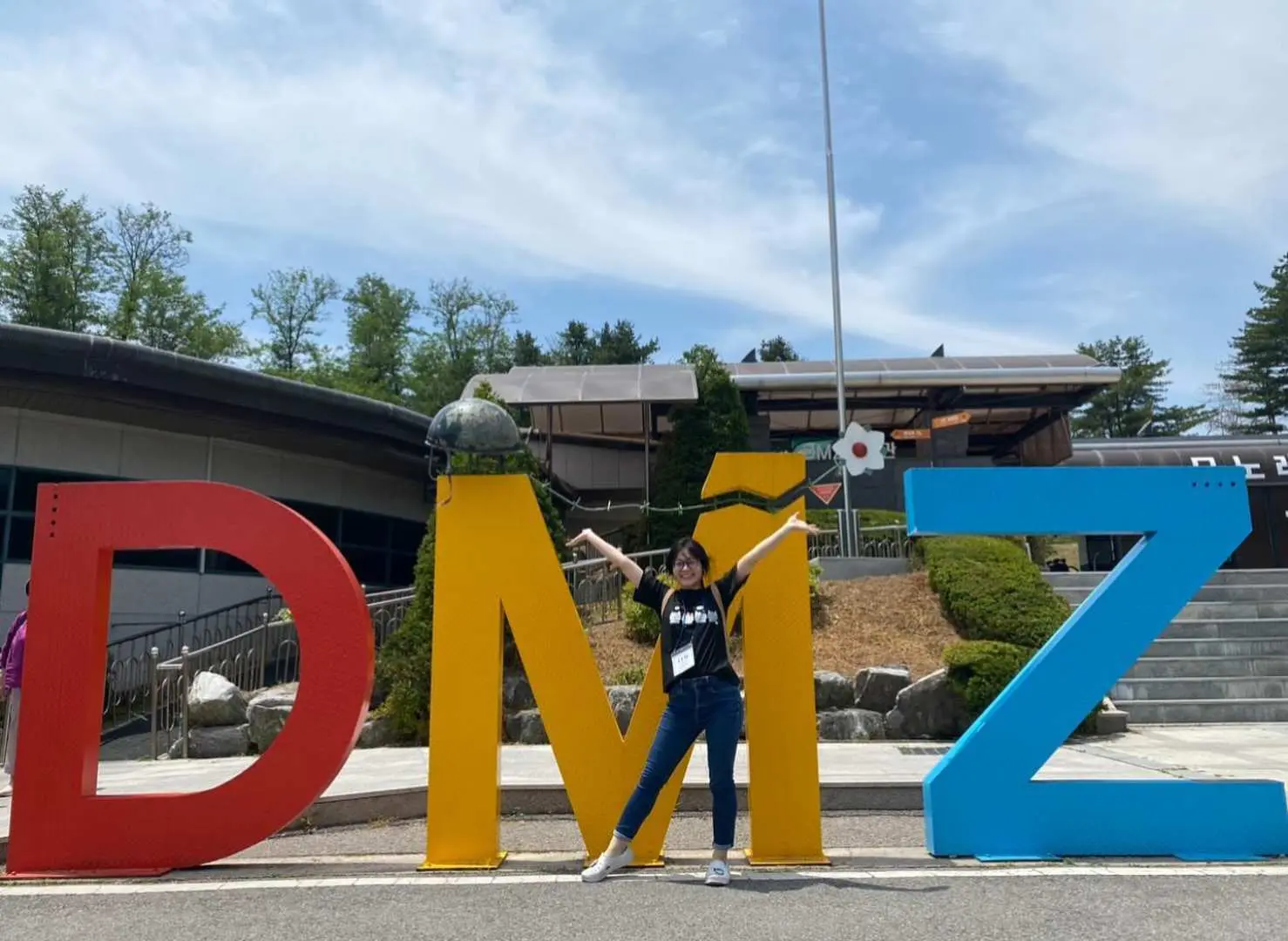 一位興奮的旅客在韓國DMZ旅遊區的巨型「DMZ」字樣地標前擺拍，享受陽光燦爛的參觀時光，捕捉獨特回憶。