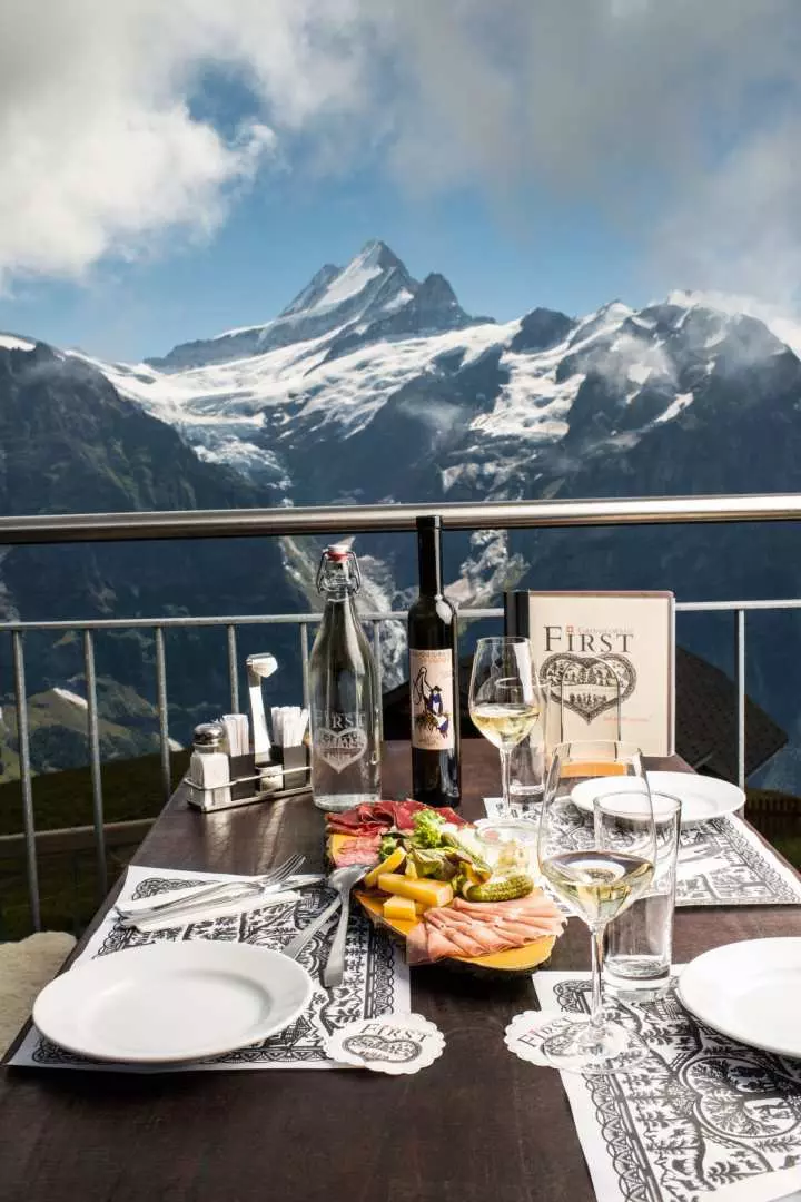A table with wine and food on the First summit restaurant terrace, with the majestic snow-capped Bernese Oberland mountains of Grindelwald, Switzerland, in the background.