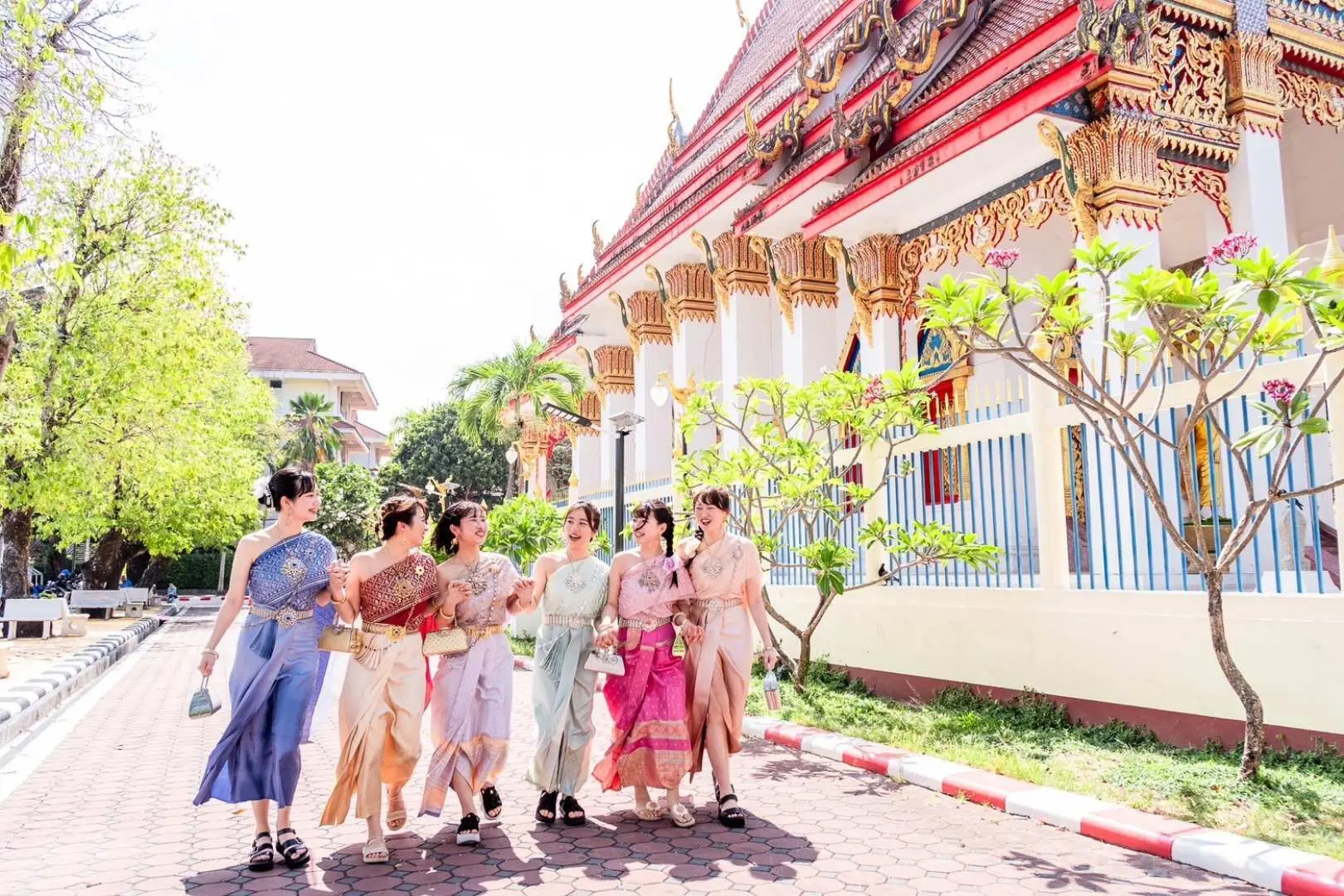 A group of women in various traditional Thai costumes happily strolling along a path next to a magnificent Phuket temple, enjoying sunshine and a cultural journey.