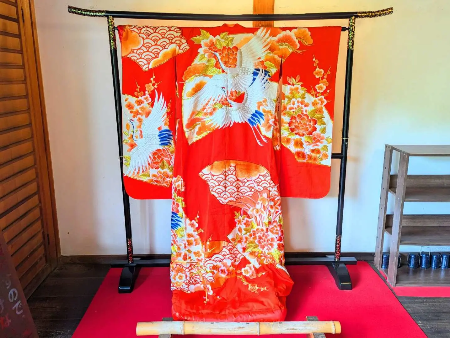 An ornate red Japanese uchikake kimono, adorned with exquisite crane and floral embroidery, displayed on a wooden rack, showcasing the artistry of traditional Japanese attire.