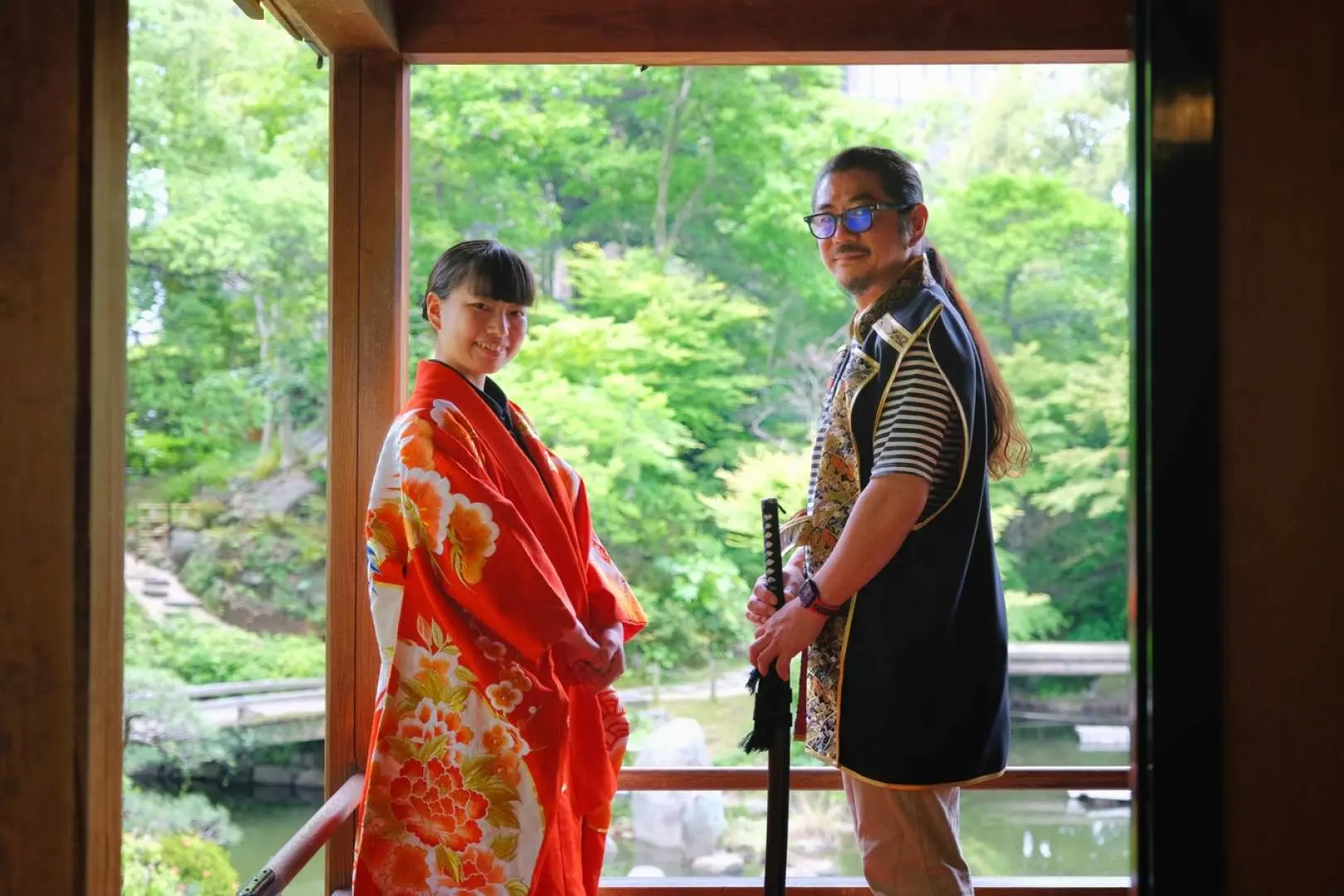 A young couple in traditional Japanese attire posing at the Kokura Castle Garden tea house, the woman in a vibrant kimono and the man with a samurai sword, against a lush garden backdrop.