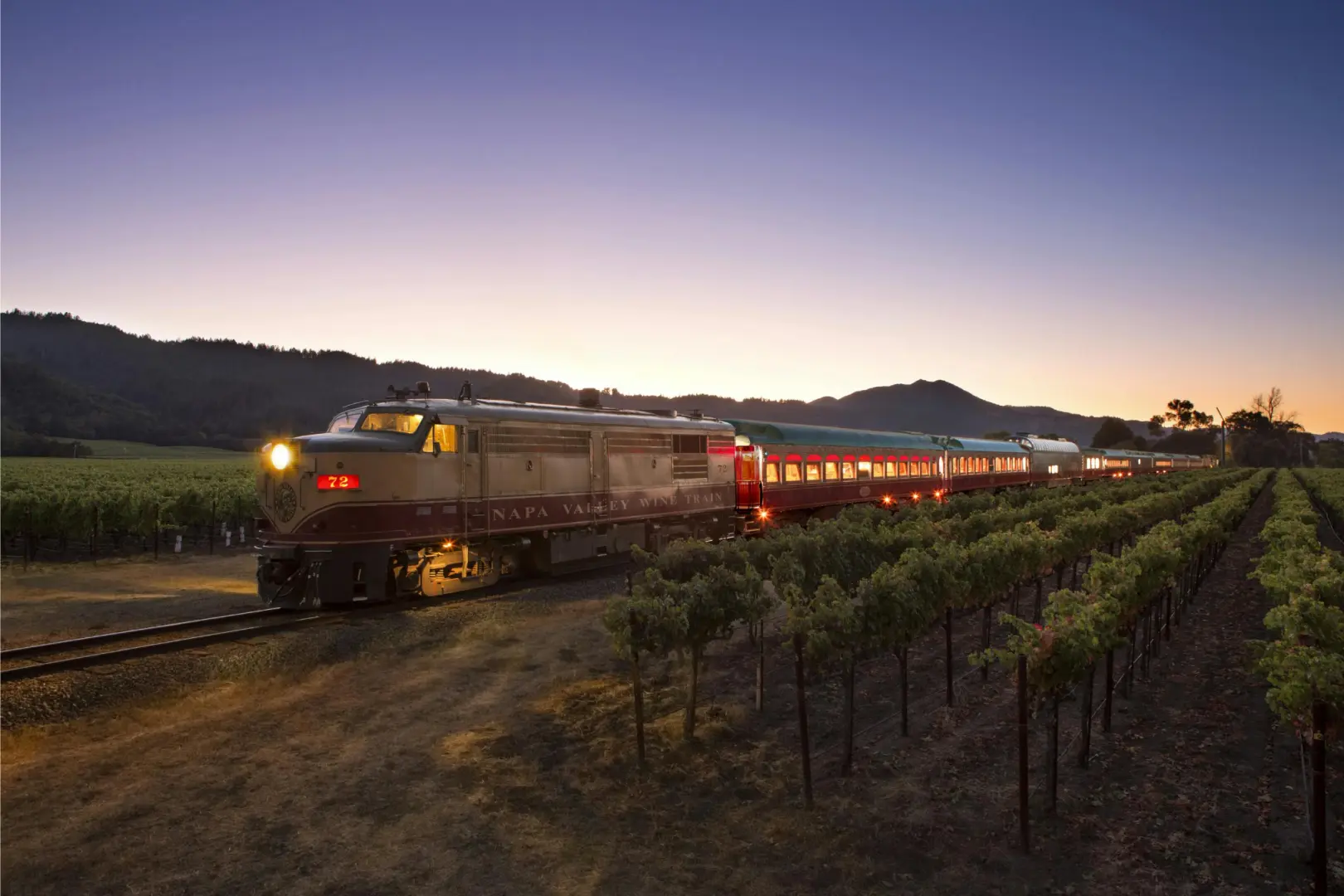 The Napa Valley Wine Train gracefully glides through lush vineyards at dusk, its lights illuminating the path, showcasing the romantic and elegant California fine dining and wine journey.