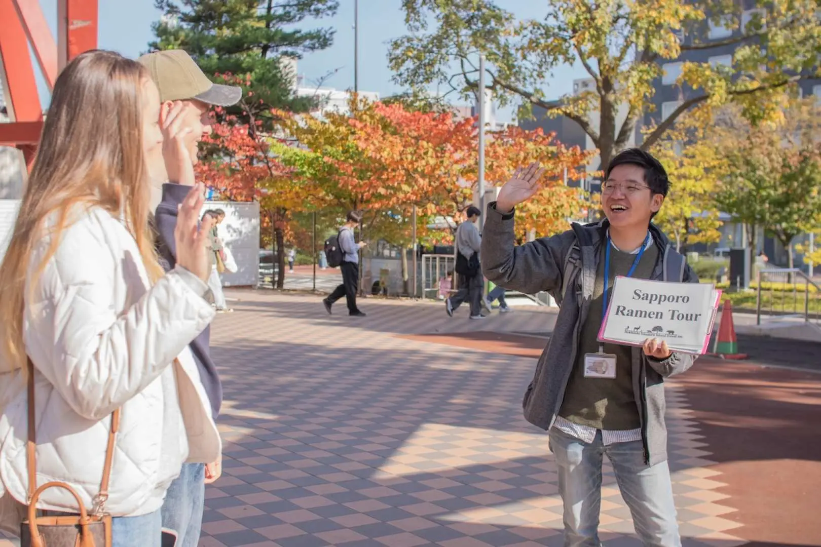 A private guide, holding a 'Sapporo Ramen Tour' sign, welcomes visitors on an autumnal Sapporo street, initiating an ultimate ramen experience combining city exploration and culinary discovery.