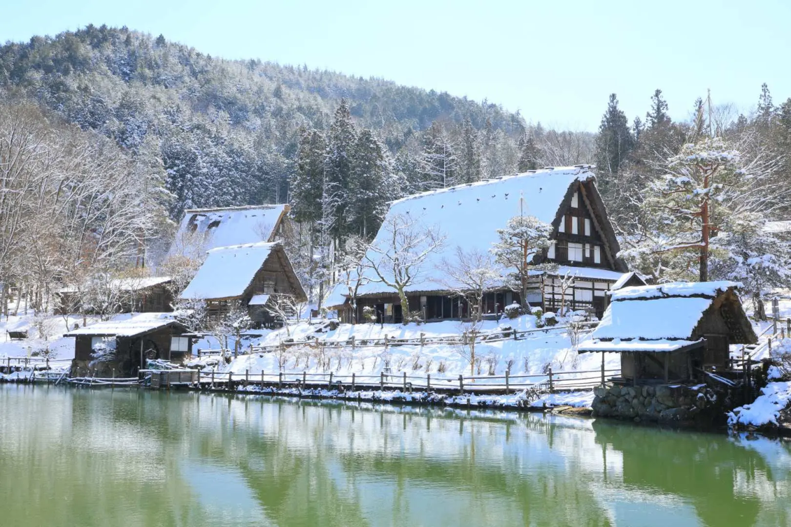 日本白川乡合掌村的冬日风景，雪白屋顶的合掌屋坐落在清澈的河畔，阳光洒落，呈现静谧而美丽的日本世界遗产风貌。