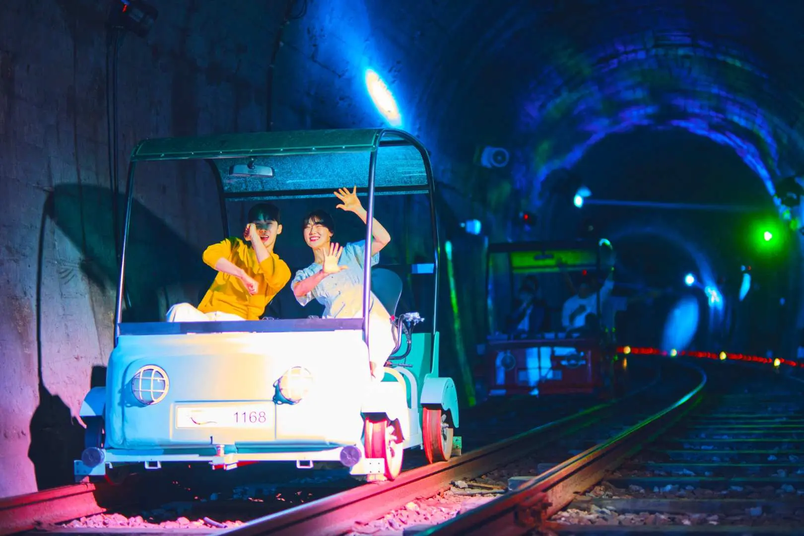 Two tourists joyfully cycle a Gangchon Rail Bike through a dazzling tunnel filled with vibrant light effects, enjoying a unique and thrilling railway experience.