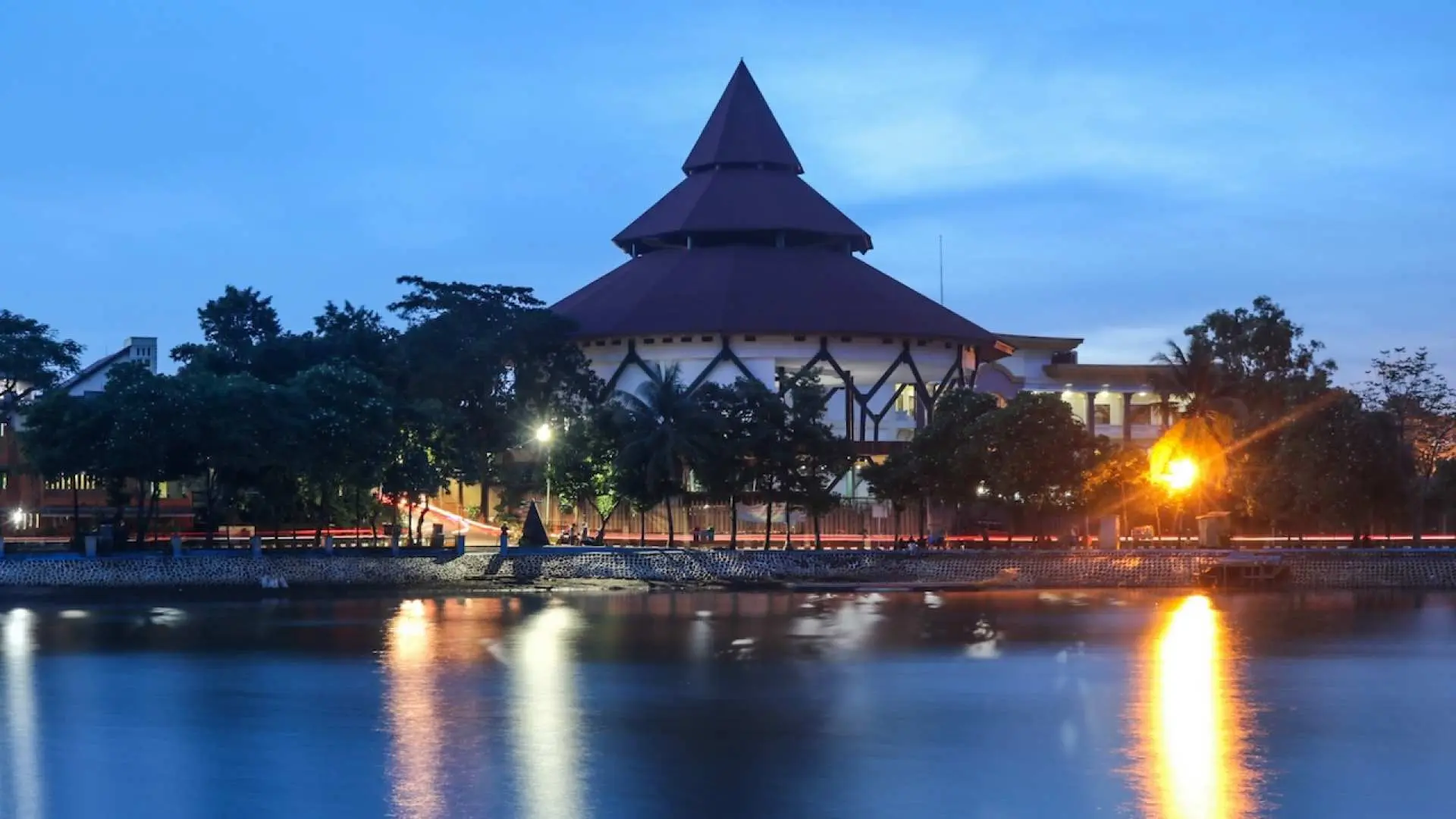 Iconic Indonesian traditional building at Taman Mini Indonesia Indah, beautifully illuminated at dusk and reflecting in the serene lake, symbolizing Indonesia's rich architectural heritage.