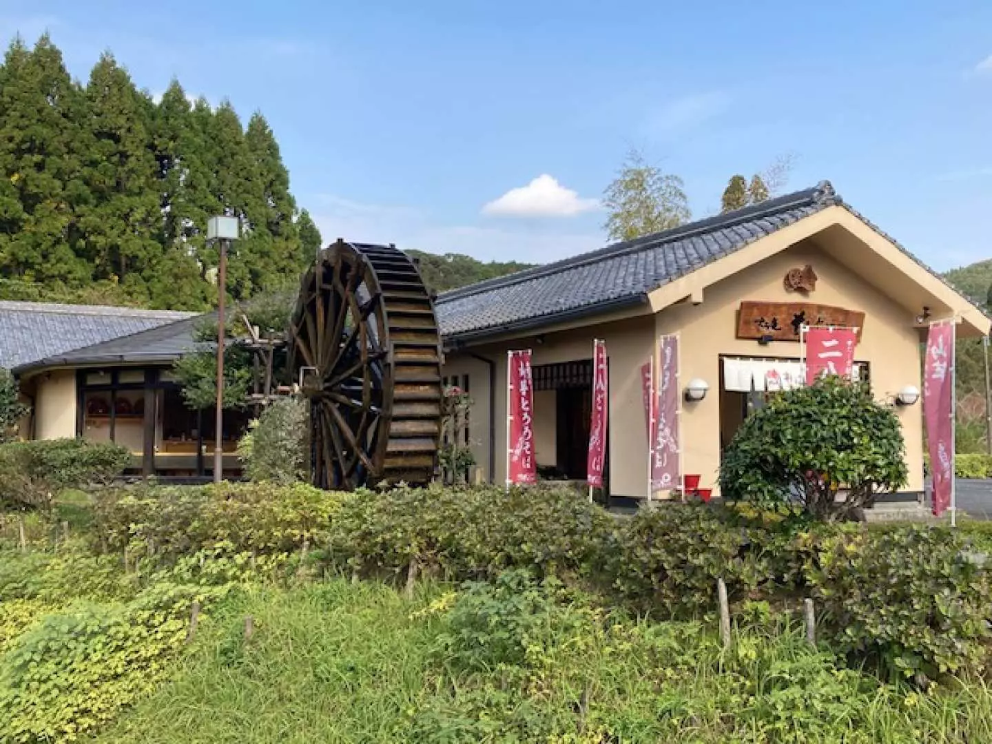 The exterior of the soba noodle making facility in Chiran, Kagoshima, featuring an antique Japanese building with a traditional water wheel, surrounded by lush natural scenery.
