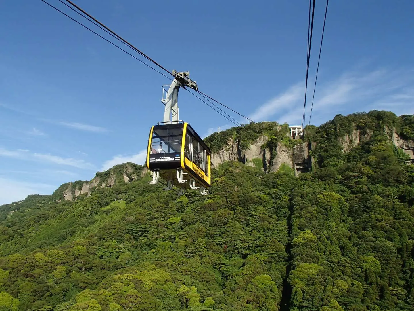 一輛黃色鋸山纜車在晴朗藍天下沿著鋼索緩緩上升，背景是千葉鋸山充滿奇石怪石的翠綠山脈。