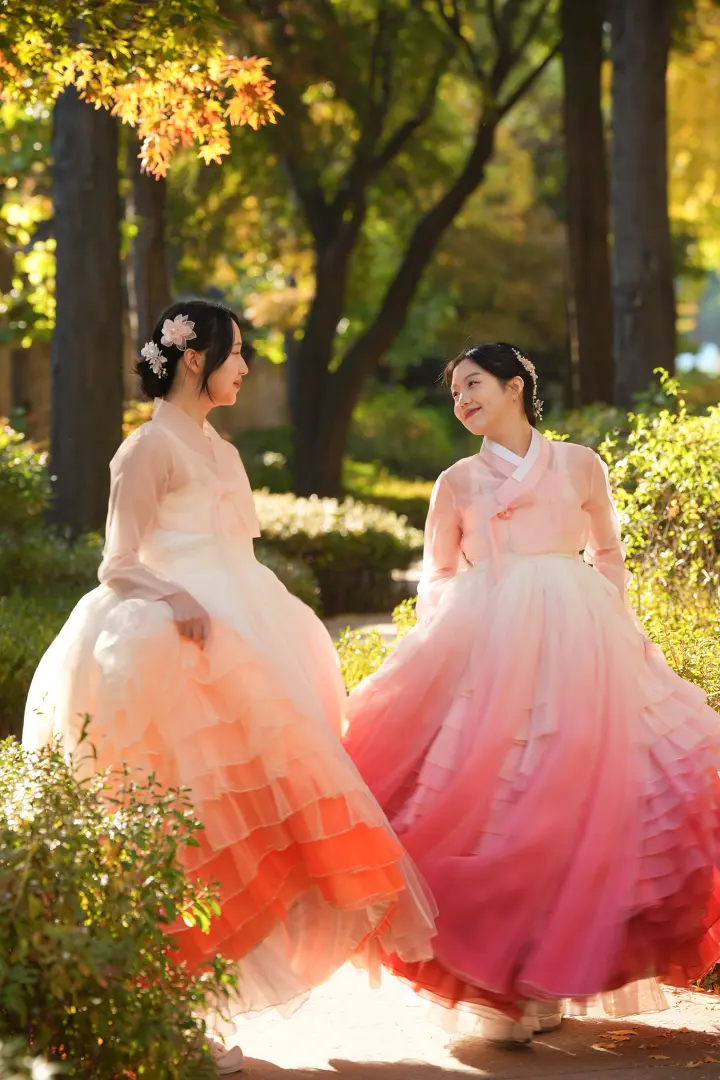 兩位女子身著色彩鮮豔、多層次漸層韓服，在陽光普照的韓國傳統庭園中歡快漫步。