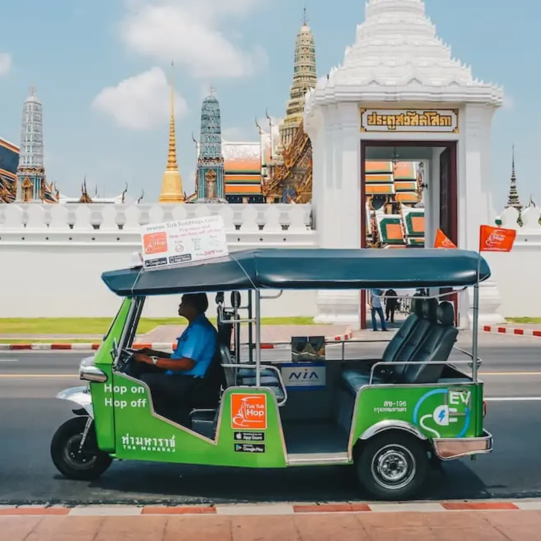 A green electric tuk-tuk parked on a Bangkok street, with the majestic Grand Palace walls in the background, offering a convenient hop-on hop-off city sightseeing experience.