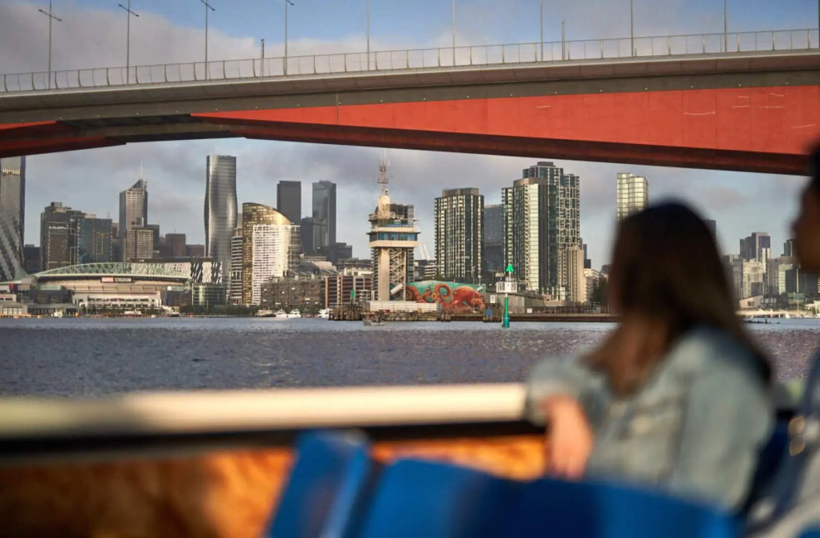 从雅拉河游船内部眺望墨尔本城市风光，高耸建筑在红色桥梁下若隐若现，游客舒适地欣赏河畔景色。