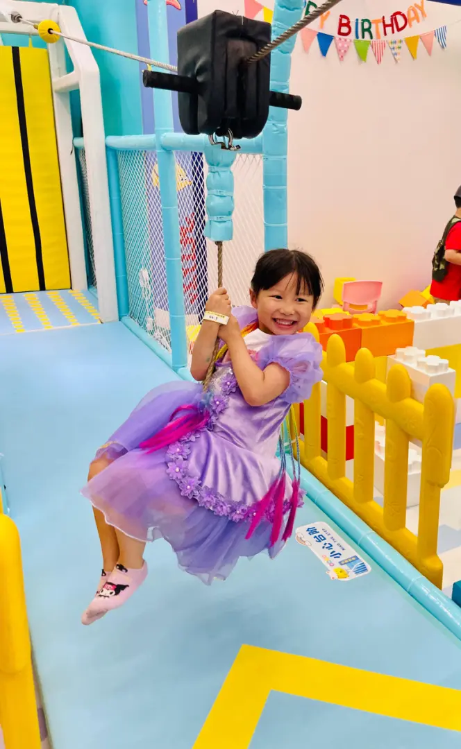 Child happily riding the indoor zip line at Sunday Island Playhouse, a young girl in a purple dress gliding through the air, enjoying a unique thrill and fun experience.