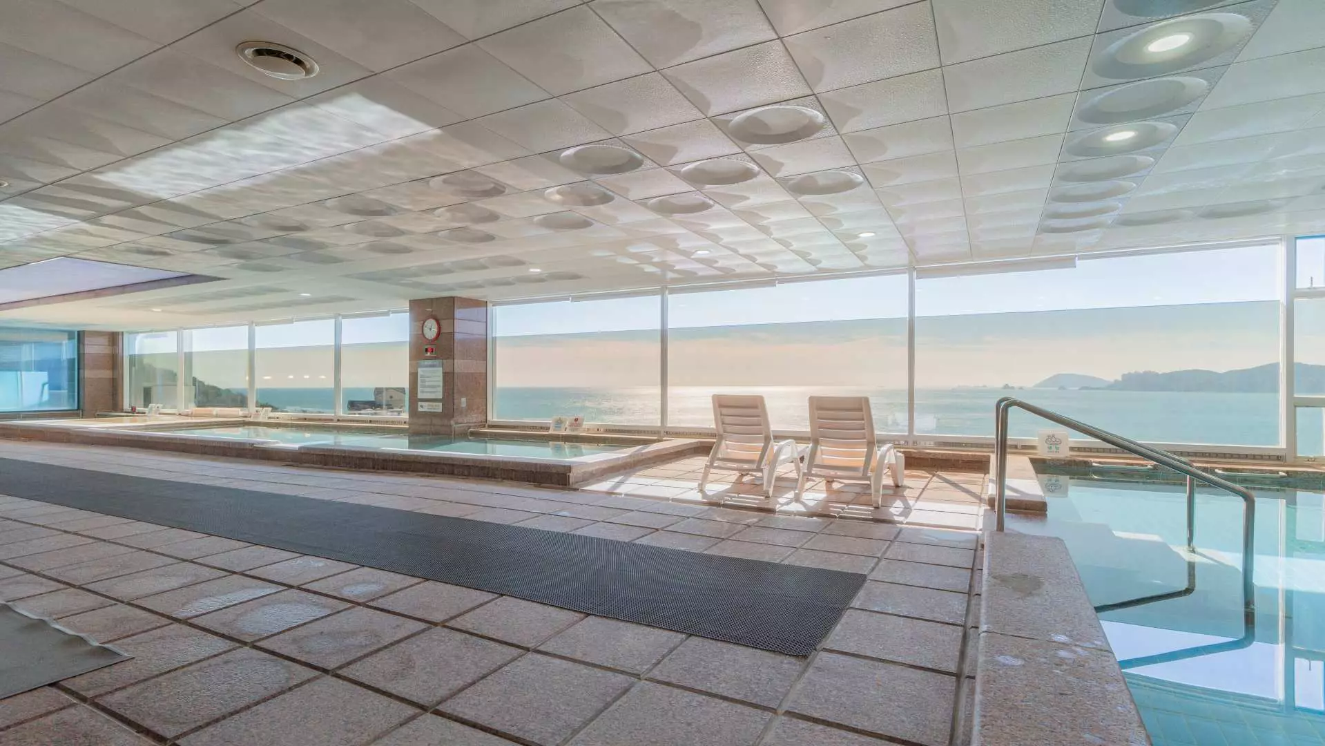 Busan Haeundae HILL SPA's hot spring area, featuring various temperature pools and lounge chairs, offering an unbeatable Haeundae sea view through large glass windows for ultimate relaxation.