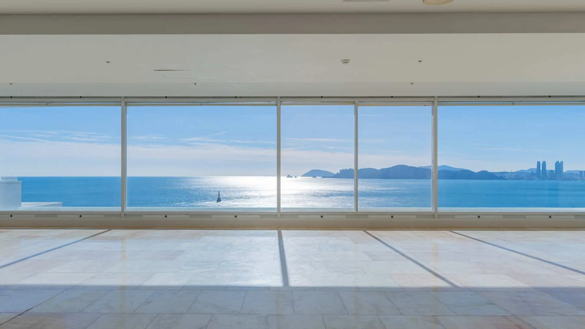 Another spacious and bright viewing area at Busan Haeundae HILL SPA, where sunlight from Haeundae fills the room through floor-to-ceiling windows, revealing expansive ocean views and cityscapes in the distance.
