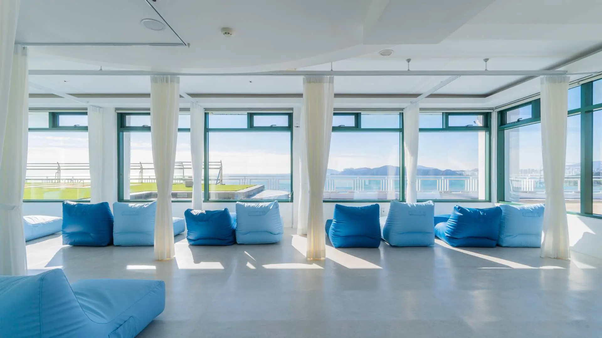 Busan Haeundae HILL SPA's spacious, bright relaxation area features comfortable blue bean bag chairs and floor-to-ceiling windows offering panoramic ocean views, perfect for unwinding.