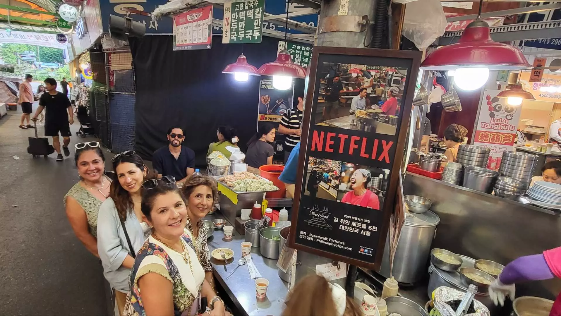 游客在首尔广藏市场充满活力的摊位间探索美食，背景可见Netflix美食纪录片标志，沉浸于韩国传统市场的文化与美食。