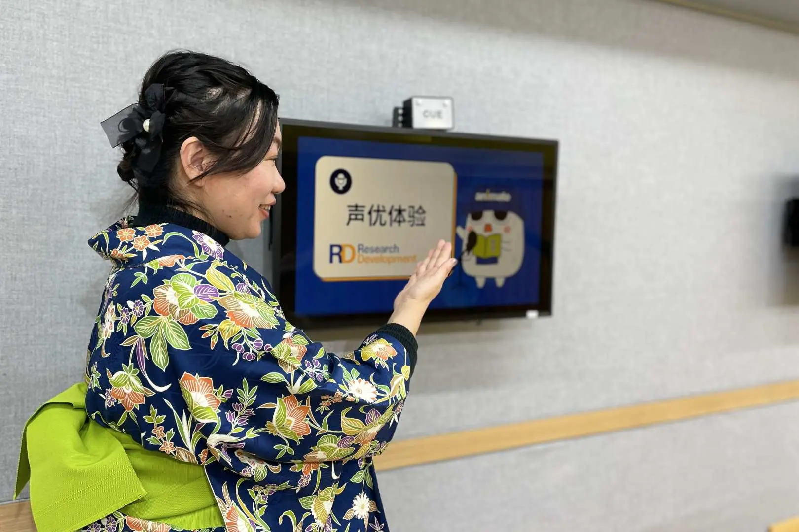 A professional voice actor instructor, dressed in an elegant kimono, introduces the exciting voice acting experience in the Tokyo Ikebukuro studio, gesturing towards a screen with the experience logo.