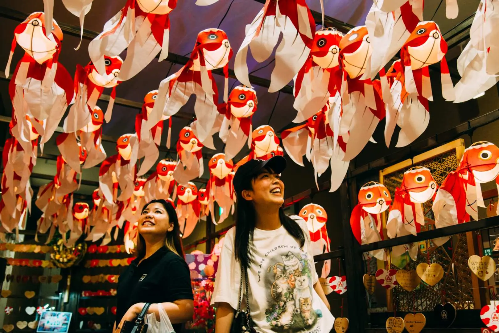Two women smiling while looking up at a canopy of red and white goldfish-shaped Japanese lanterns at the Okinawa Goldfish Museum.