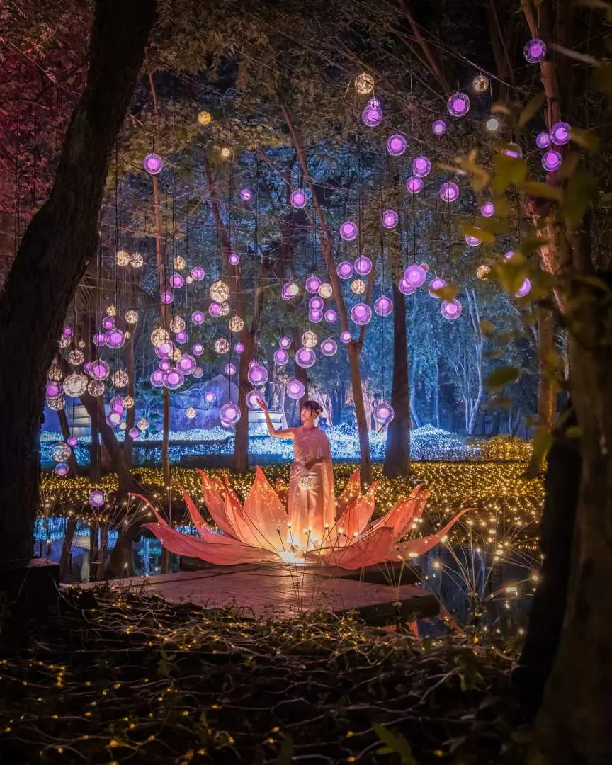 At Nasatta Park Light Festival in Thailand, visitors sit in a giant glowing pink lotus flower on water, surrounded by purple floating light orbs suspended among trees, creating a dreamlike scene.