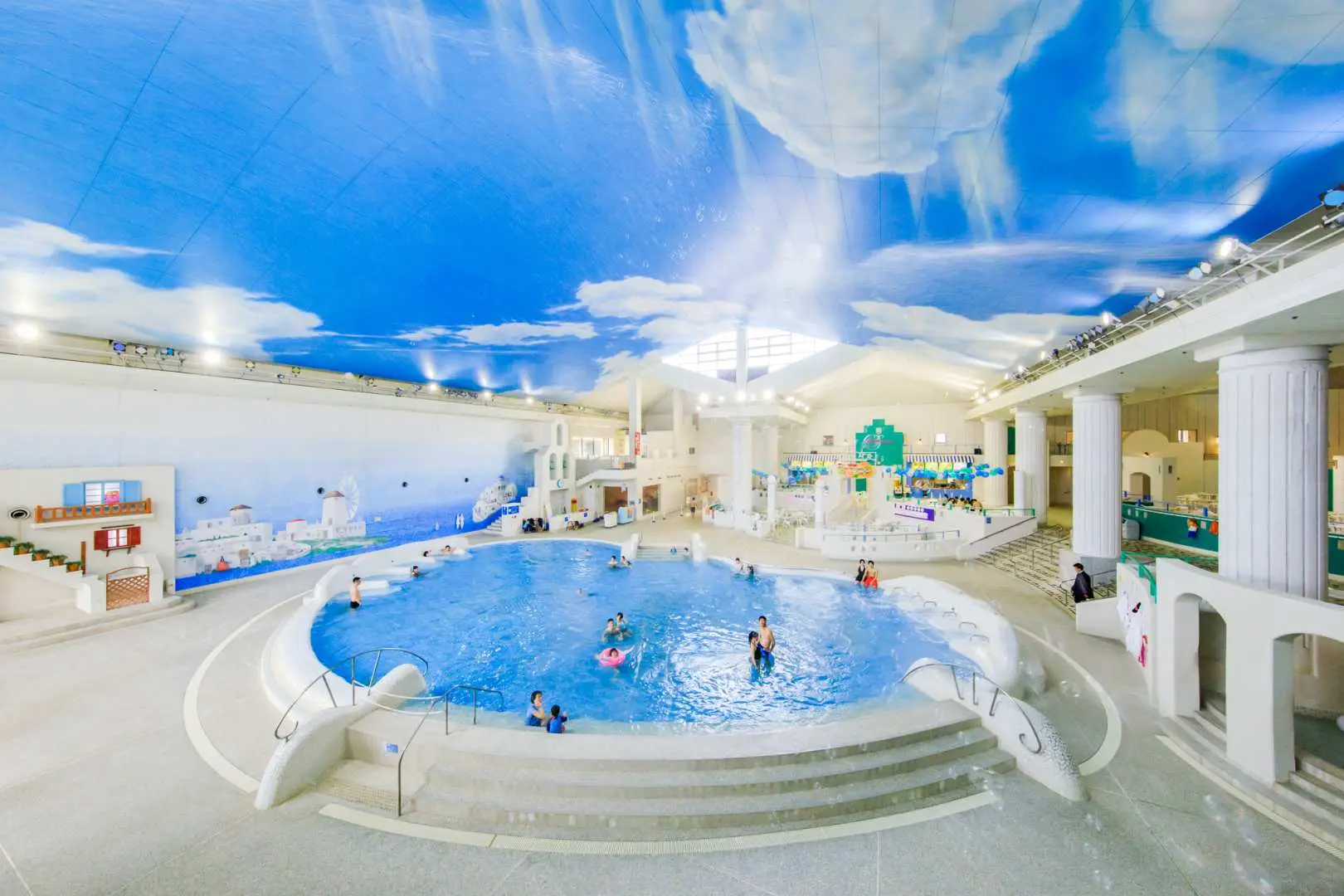 箱根小涌園ユネッサンの屋内メインプール。天井には青空と雲が描かれ、多くの利用者が水中で遊泳し、全天候型ウォーターアクティビティを楽しんでいます。