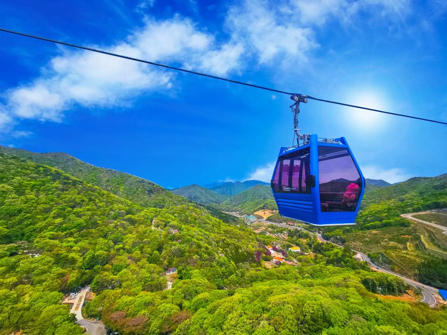 A blue Geoje Panorama Cable Car cabin slowly ascends over lush green mountains under a clear blue sky, offering breathtaking views where mountains meet the sea, a visual delight of the Geoje day trip.