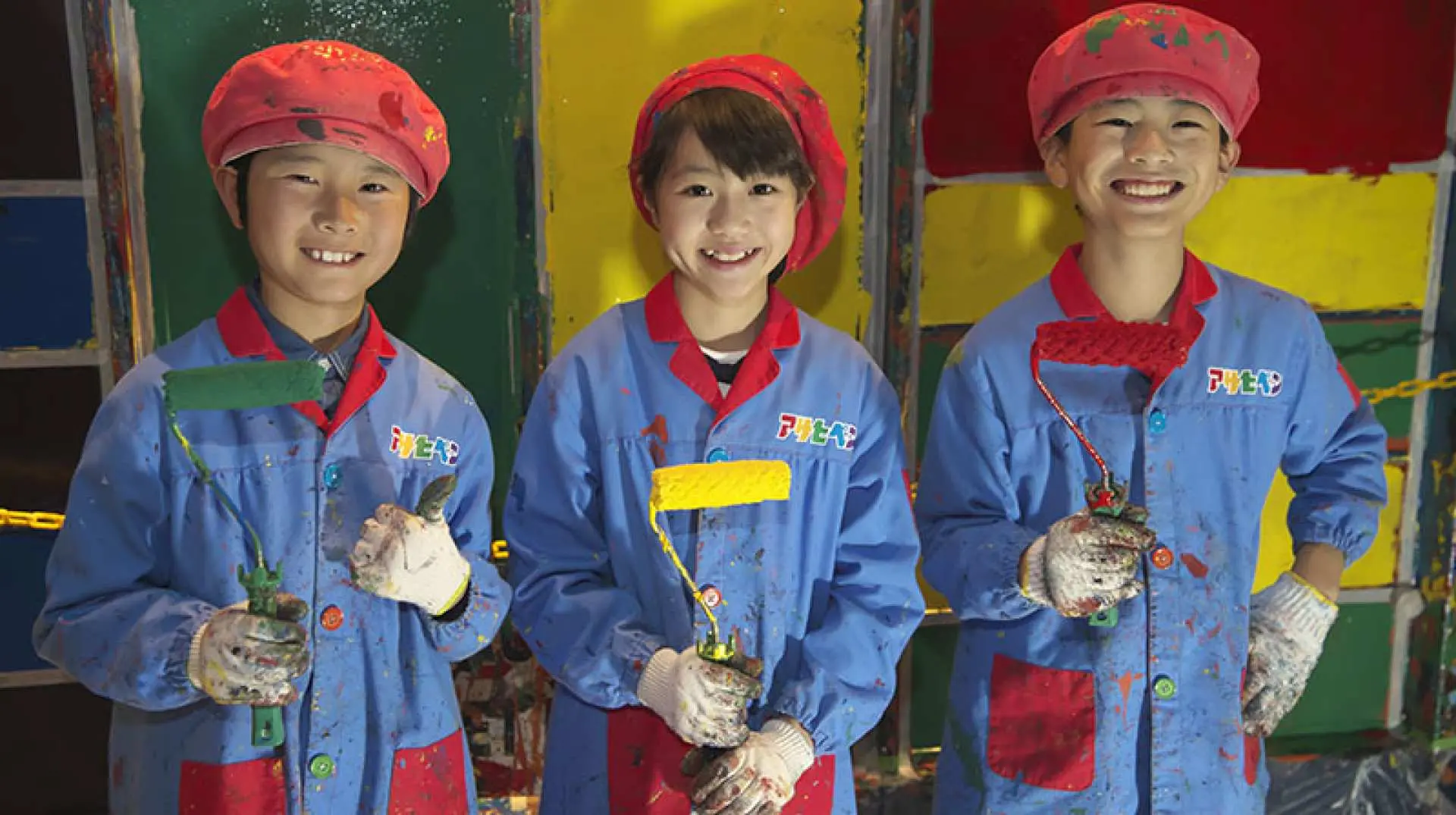 キッザニア甲子園で、青い作業服と赤いベレー帽を身につけた3人の子供たちが、ローラーを手に壁のペイントを楽しく体験し、芸術的才能を発揮しています。
