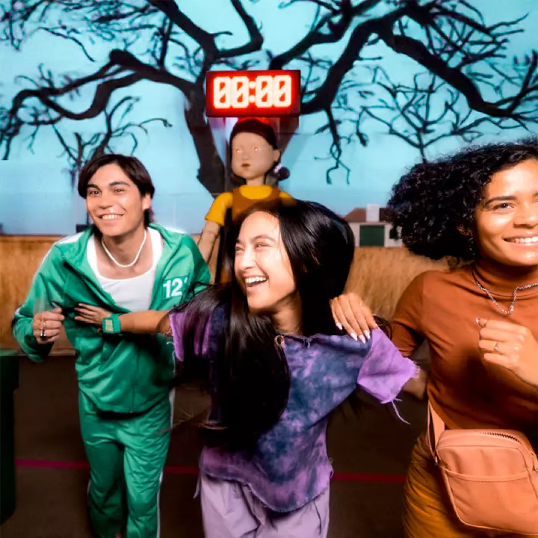 Three excited participants running during the Squid Game 'Red Light, Green Light' challenge at Sydney Luna Park, with the Young-hee doll and timer in the background, creating a tense atmosphere.