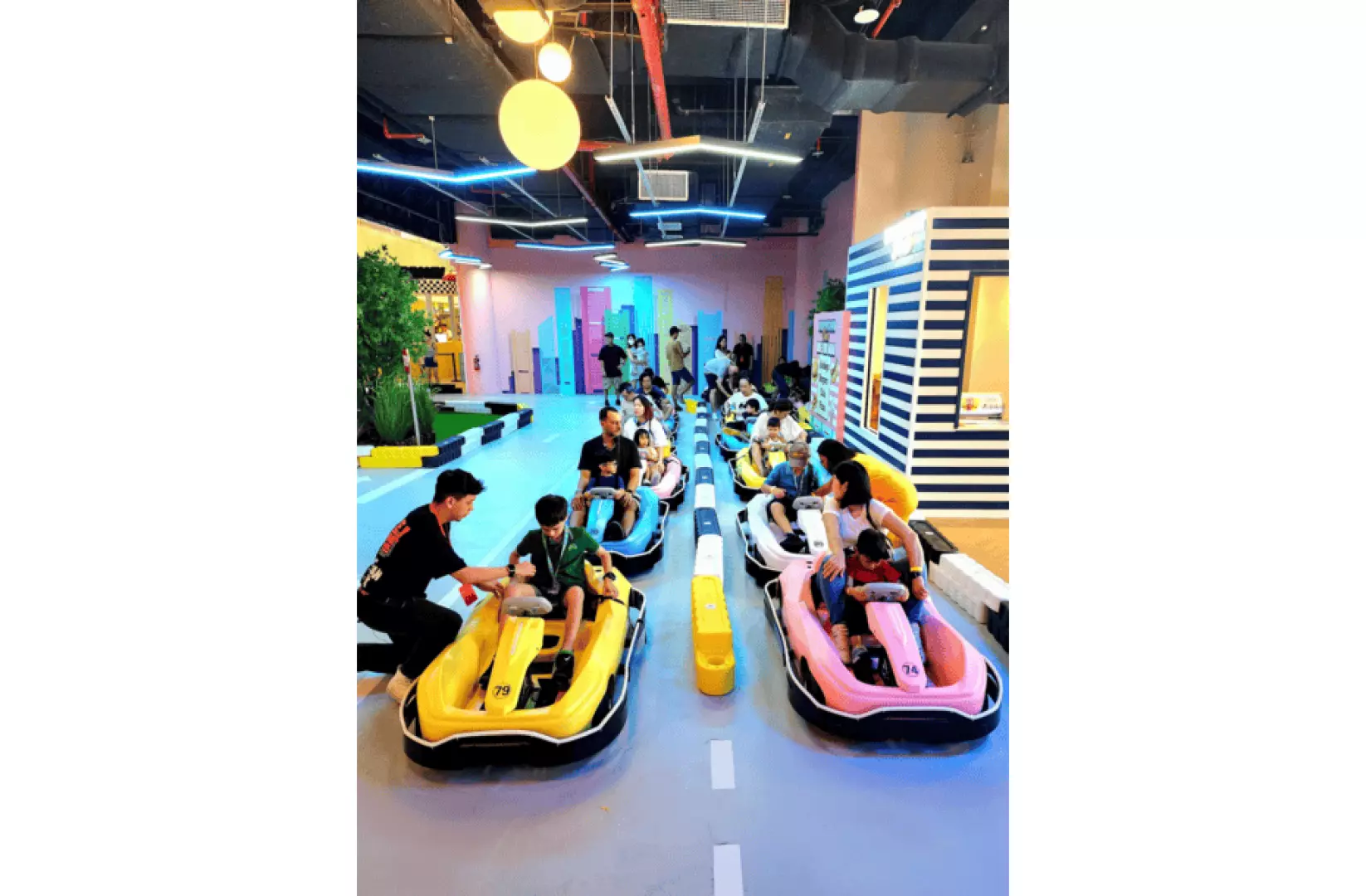 Children and parents enjoy driving bright yellow and pink VroomCars on the indoor city streets of VroomTown, Singapore's first interactive kids' driving attraction.