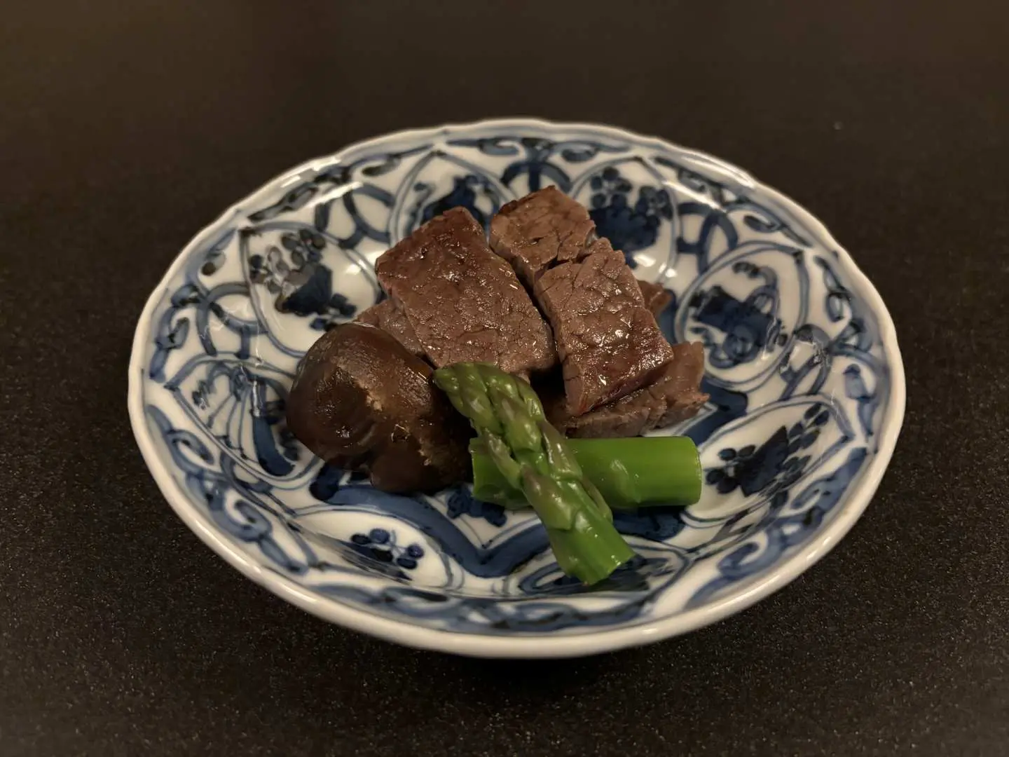 大阪味吉兆懷石料理中的一道精緻肉類小碟，深色肉塊搭配鮮綠蘆筍，盛裝於古典紋飾瓷盤中，呈現濃郁「旨味」。