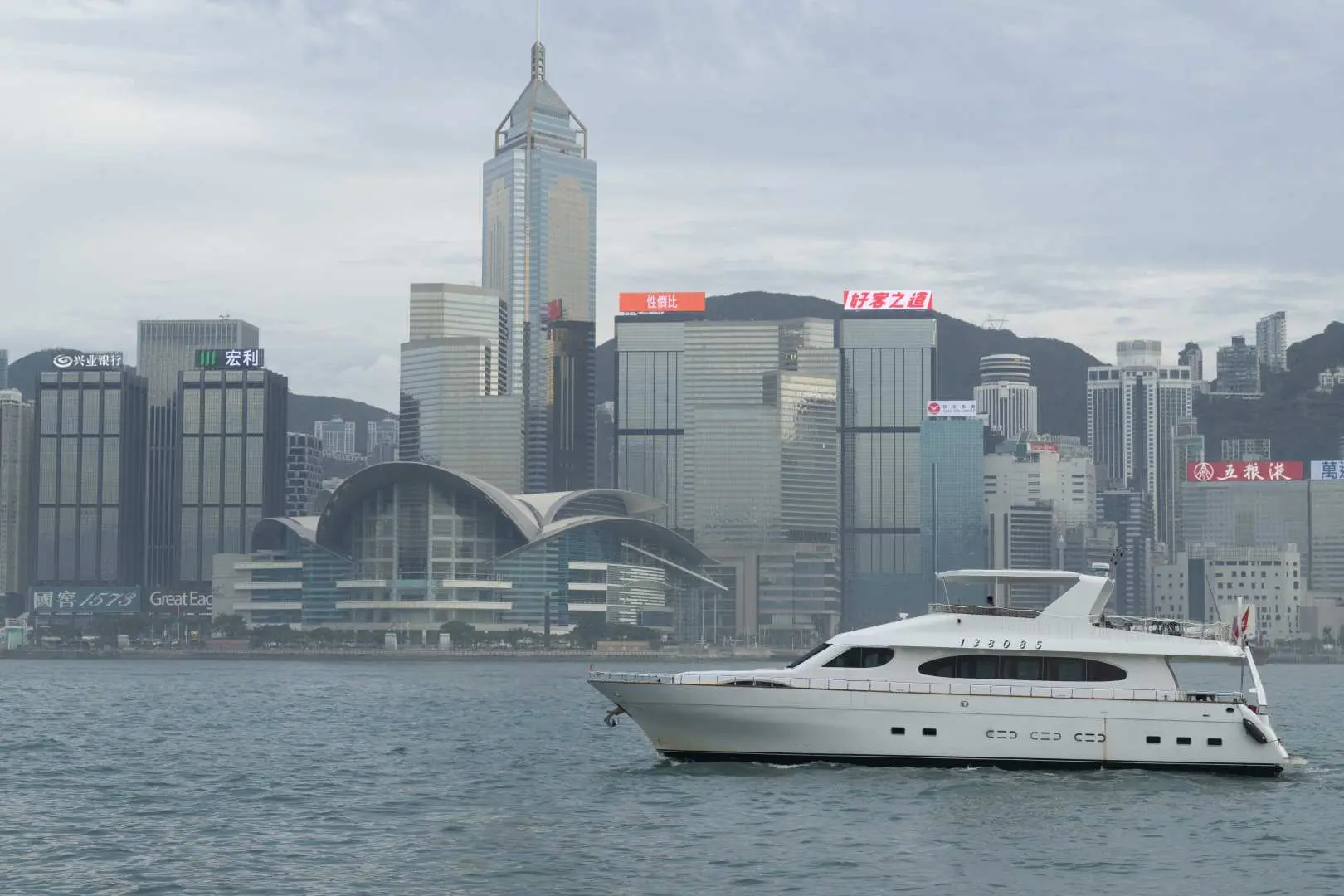 一艘白色豪华游艇在维多利亚港上航行，背景是香港中环标志性的建筑群和城市天际线。