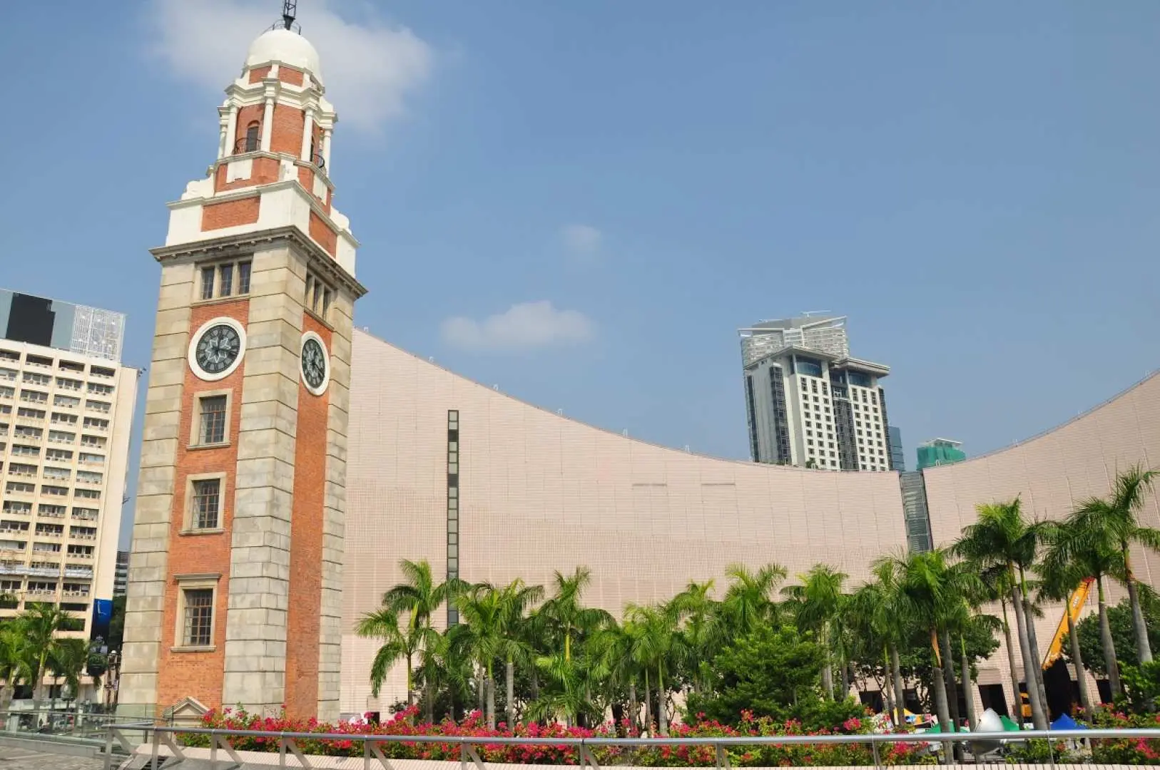 The iconic Tsim Sha Tsui Clock Tower, a historic red-brick and white-stone structure, stands tall under a blue sky, surrounded by green trees, a nostalgic must-visit landmark in Hong Kong.