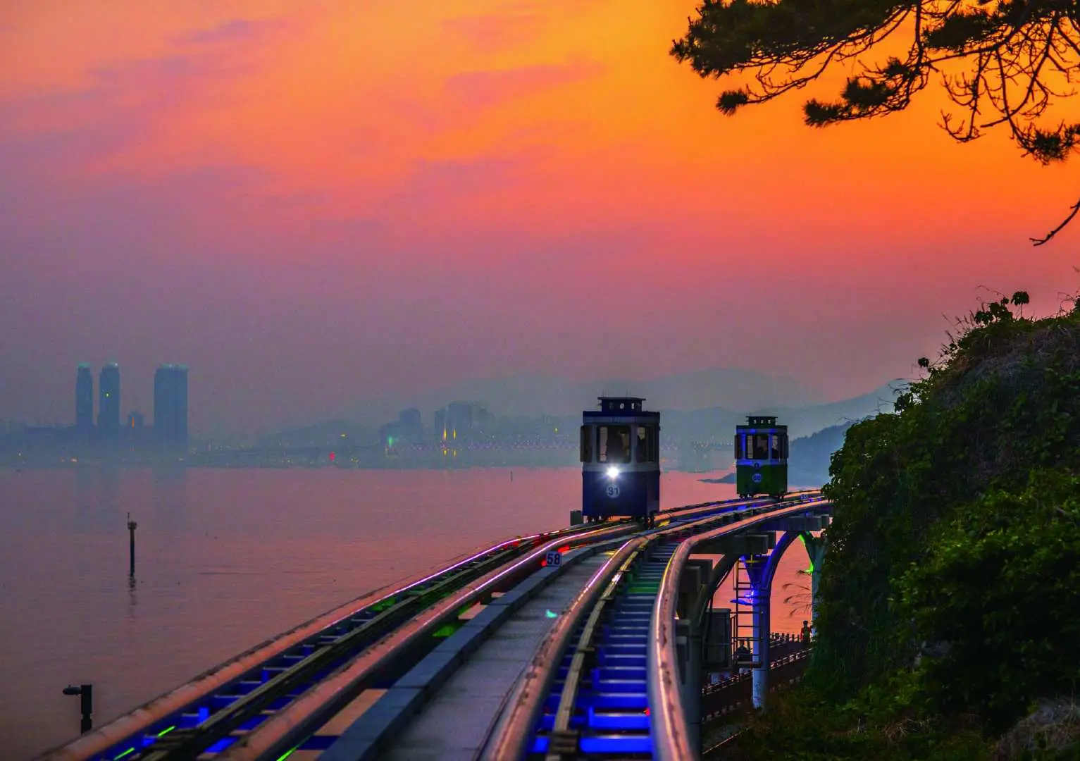 釜山天空膠囊在夕陽餘暉下沿著海岸線緩緩行駛，粉橙色的天空倒映在水面，呈現出迷人的海濱日落景色。