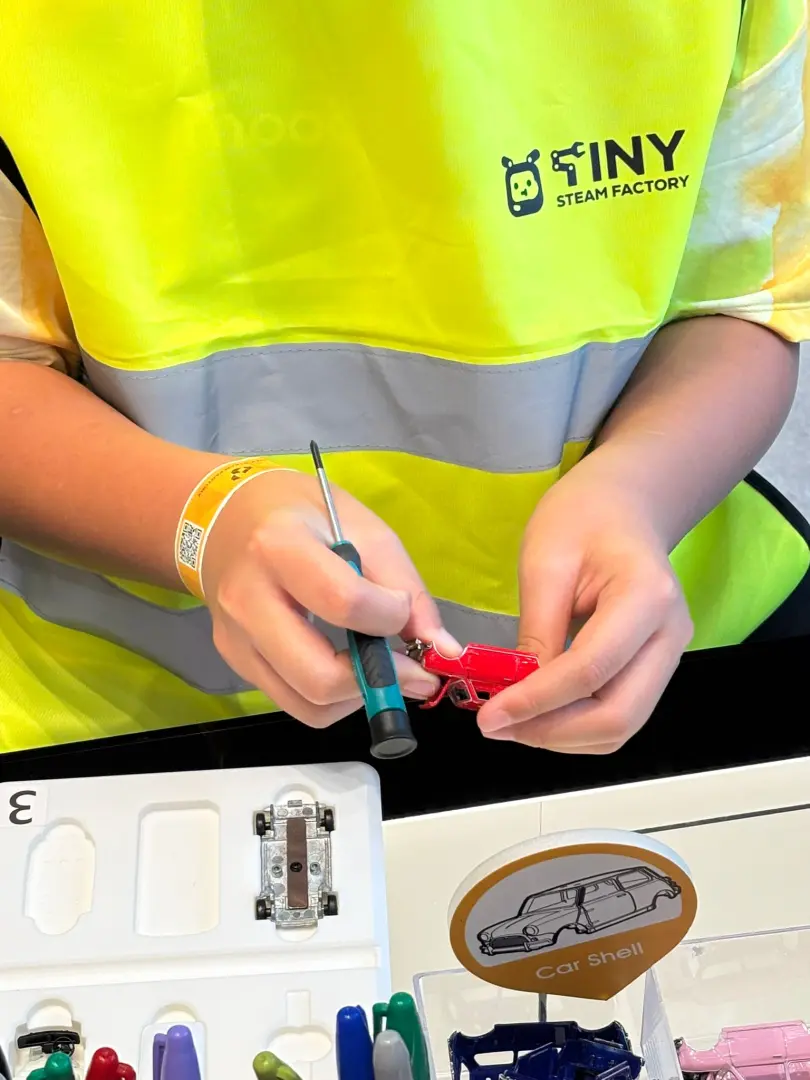 A participant wearing a Tiny Steam Factory vest intently assembles a red mini-car with a screwdriver, demonstrating the practical process of the DIY manual car assembly workshop.