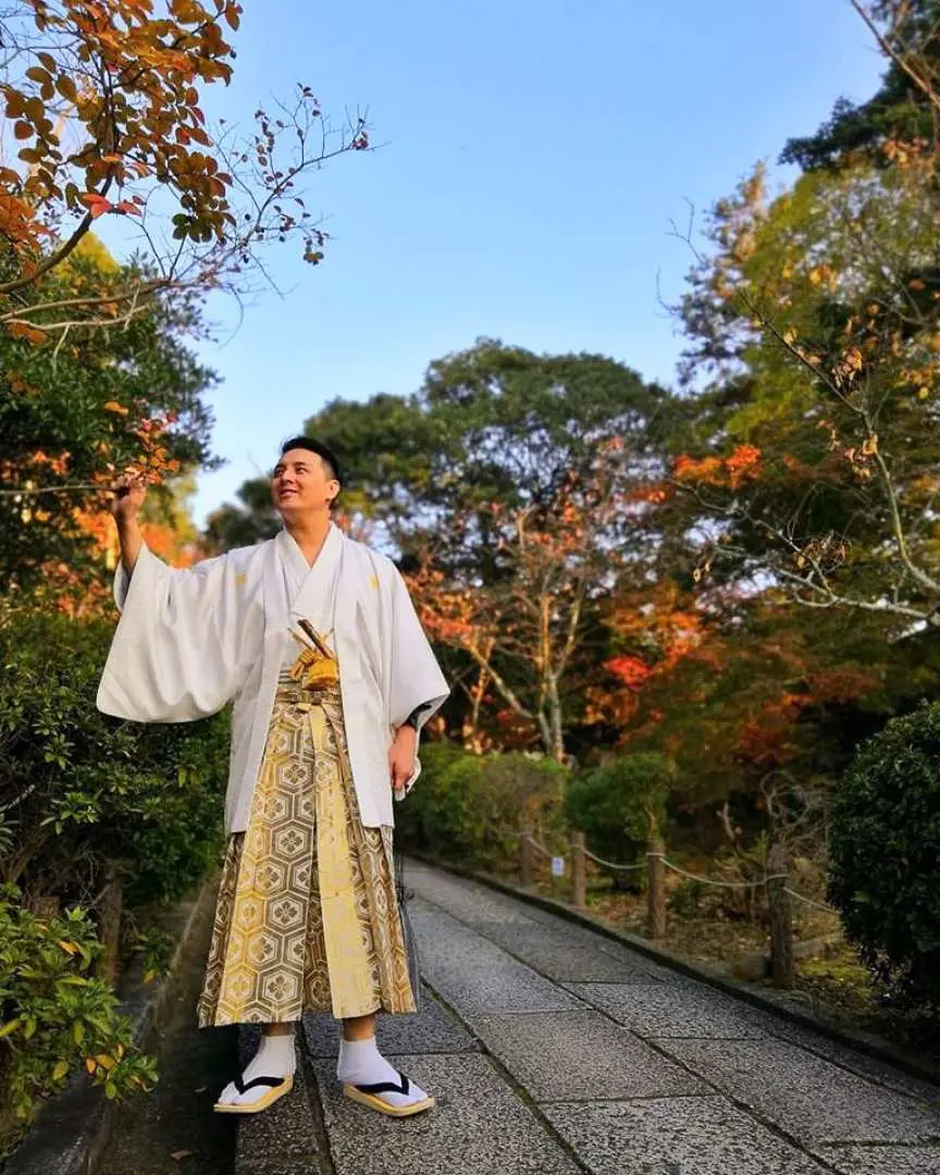 京都祇園の紅葉を背景に、白い羽織と柄袴を着た男性がポーズをとり、HANA華の男性用着物レンタルを体験しています。