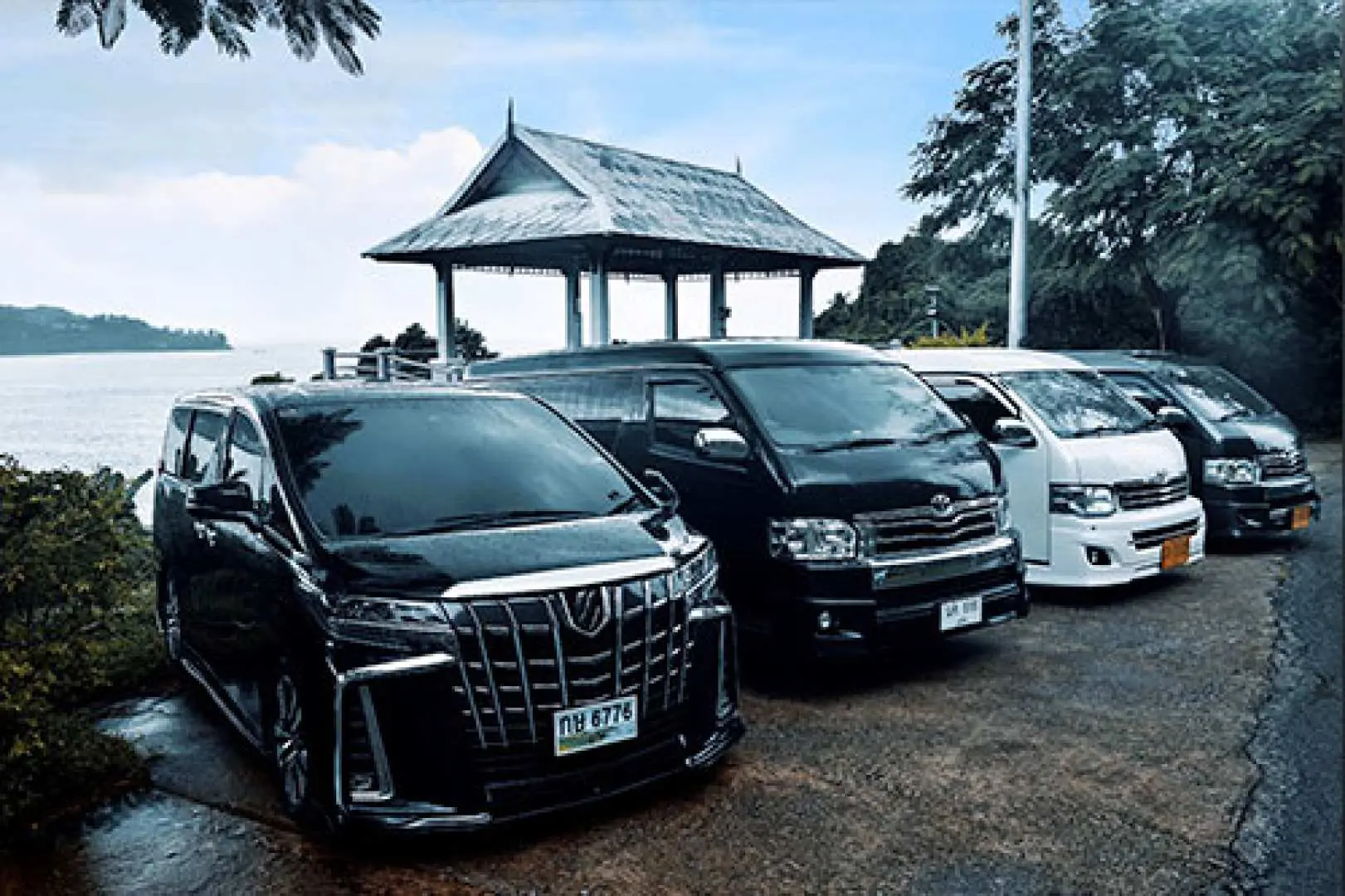 プーケットの海辺にあるタイ風ガゼボの横に駐車された複数の高級プライベートバン。空港送迎や島内観光に信頼性の高い車両を提供します。