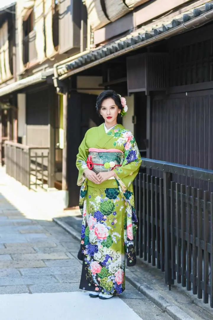 女子身著亮綠色高級打掛和服，頭戴優雅花飾，在京都充滿古樸風情的傳統街道上，由專業攝影師拍攝全身照。