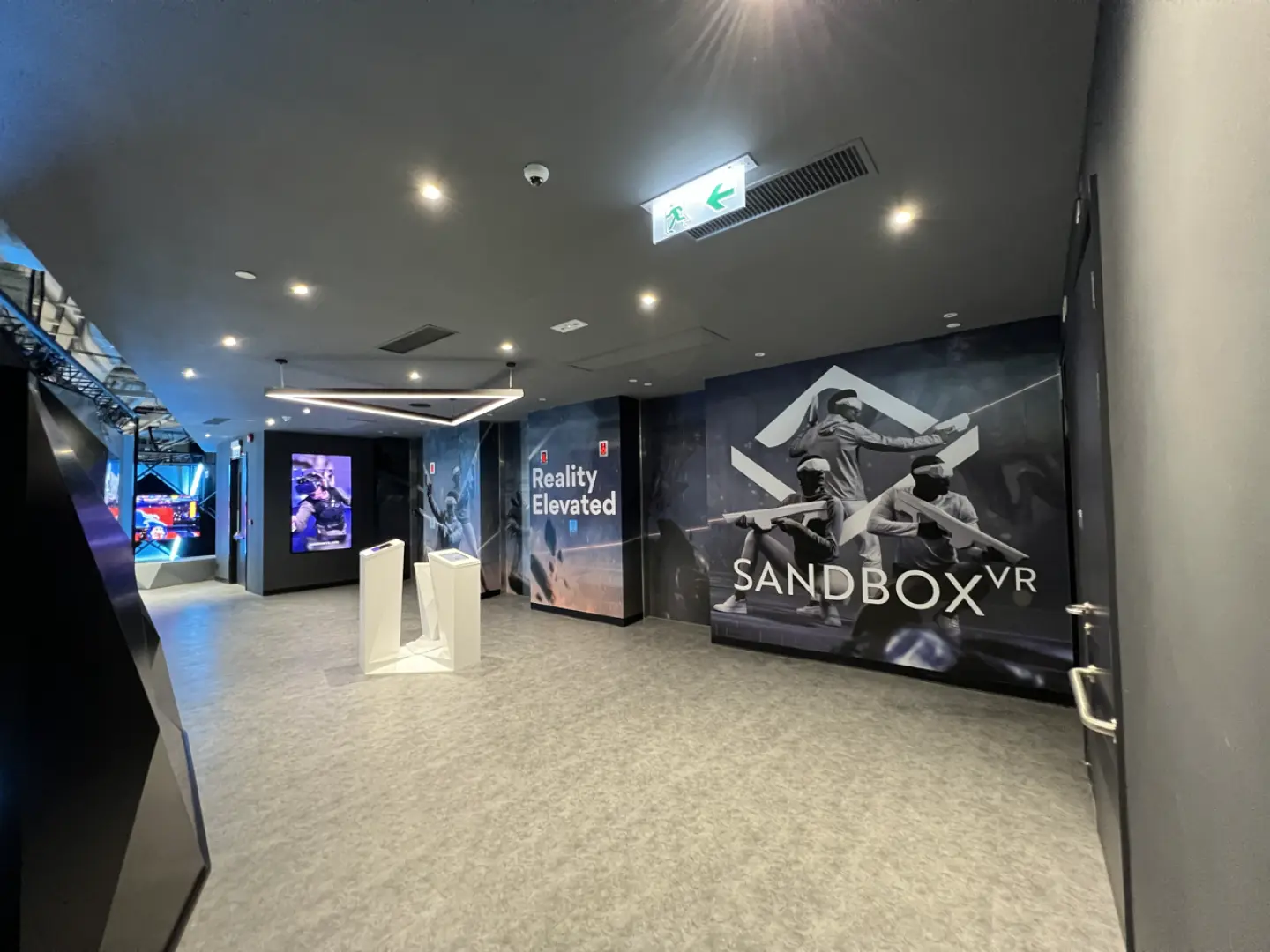 香港Sandbox VR尖沙咀或銅鑼灣店內現代寬敞嘅走廊環境，牆面掛有「Reality Elevated」標語同遊戲角色宣傳海報，引導顧客進入VR世界。