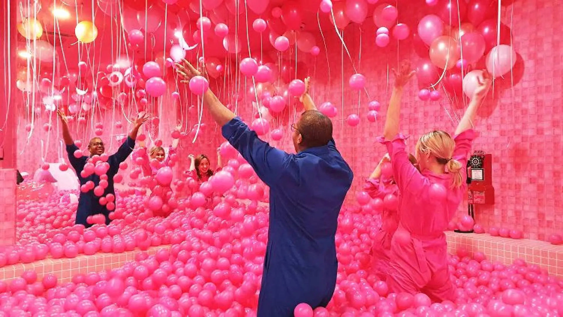 Visitors immerse themselves in a vibrant pink ball pit, raising their arms amidst countless pink balloons hanging from the ceiling, enjoying the energetic atmosphere at Seoul's Insa-dong Color Pool Museum.